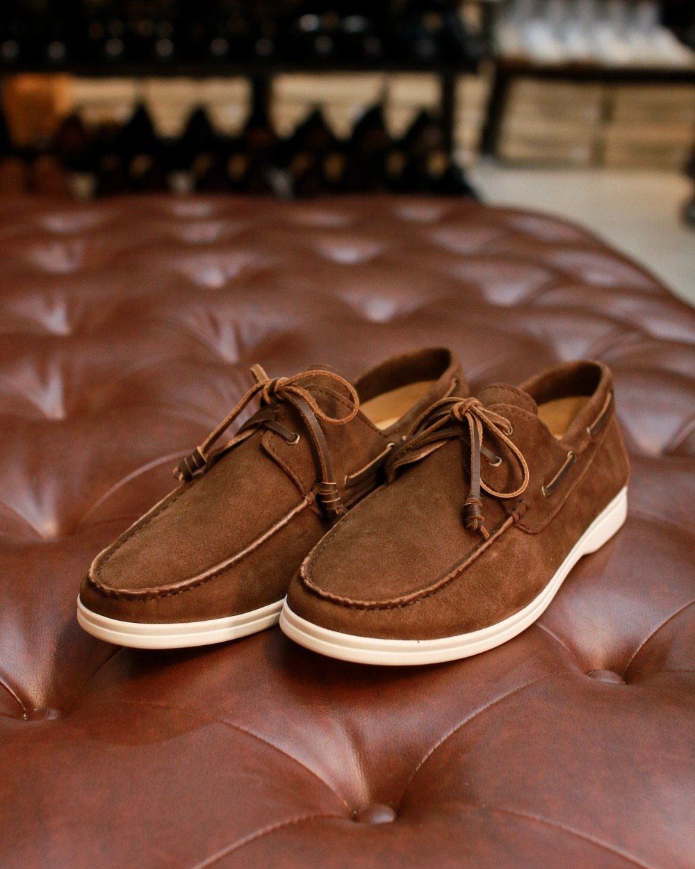 Brown suede boat shoes with white soles resting on a tufted brown leather surface.