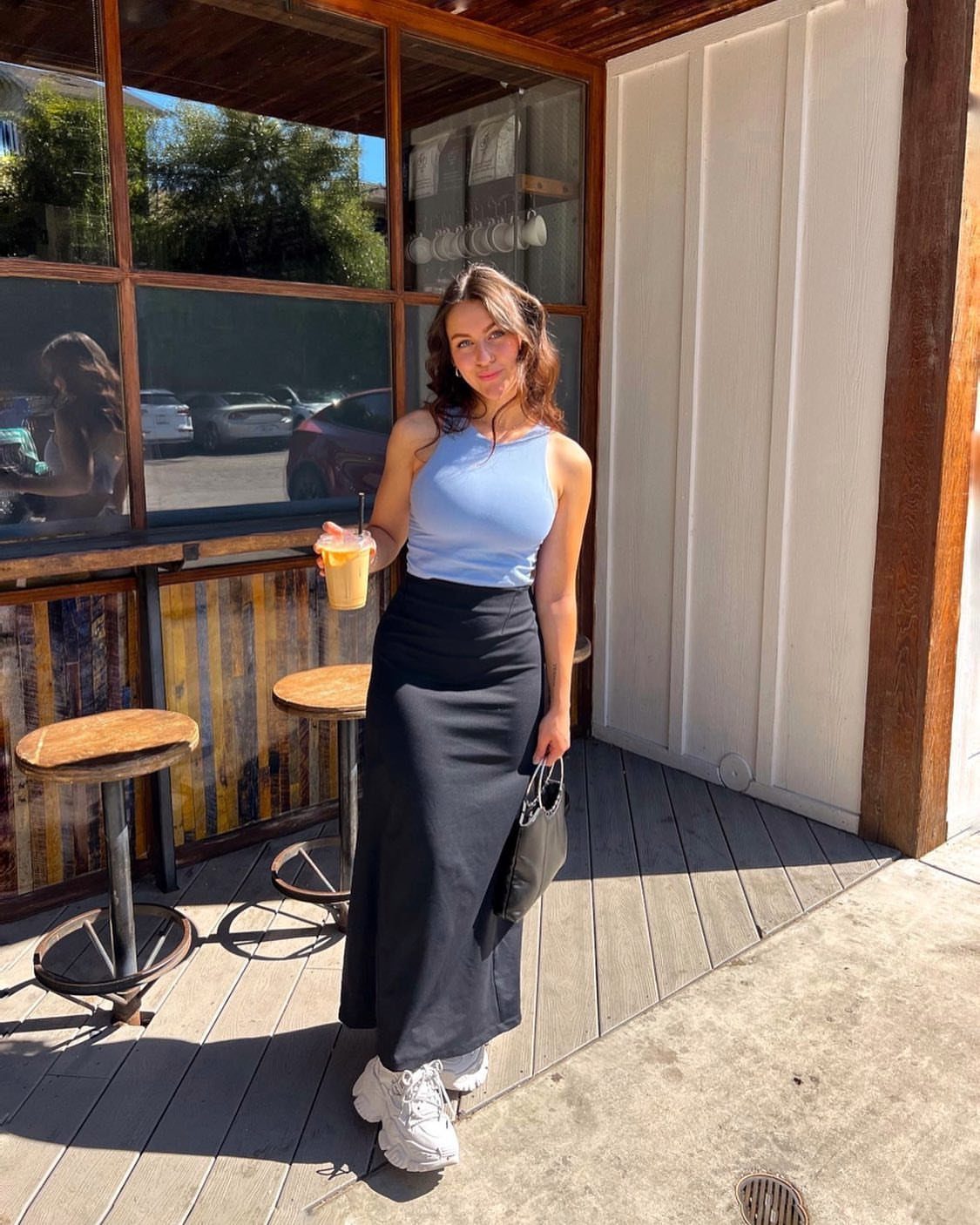 A woman wearing a long black skirt and chunky white sneakers standing on a wooden deck.