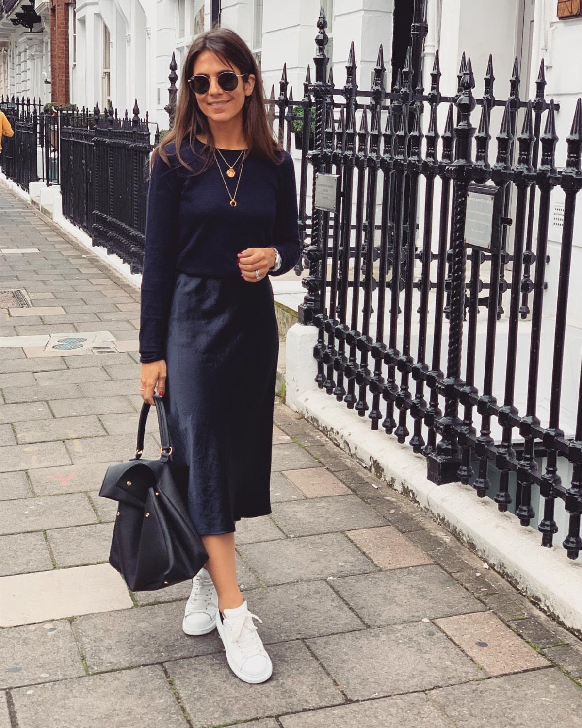 A woman in a black satin skirt and white sneakers posing on a city sidewalk.