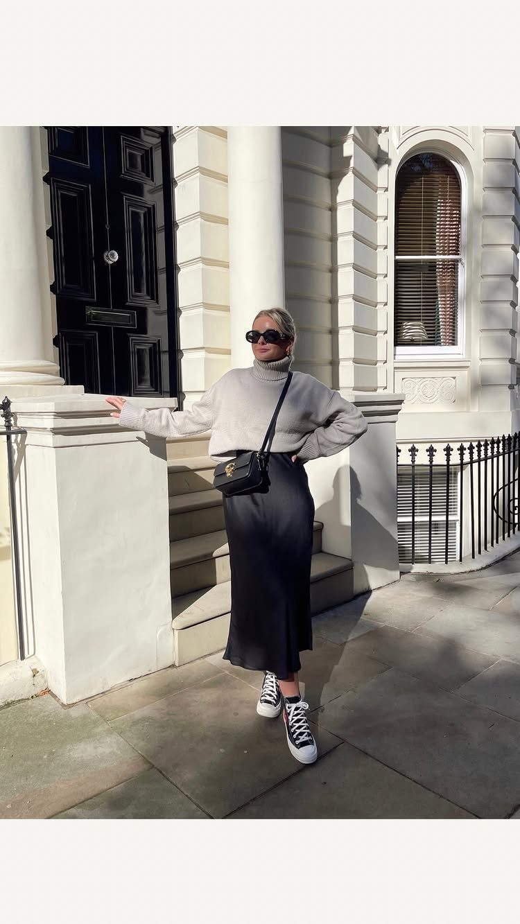 Woman wearing a grey sweater, long black skirt, and black and white sneakers standing outdoors.