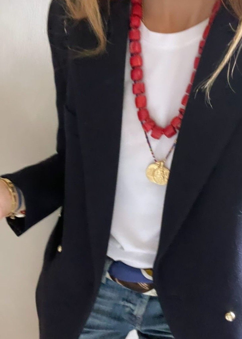 Person wearing a navy blazer, white t-shirt, and layered red and gold necklaces.