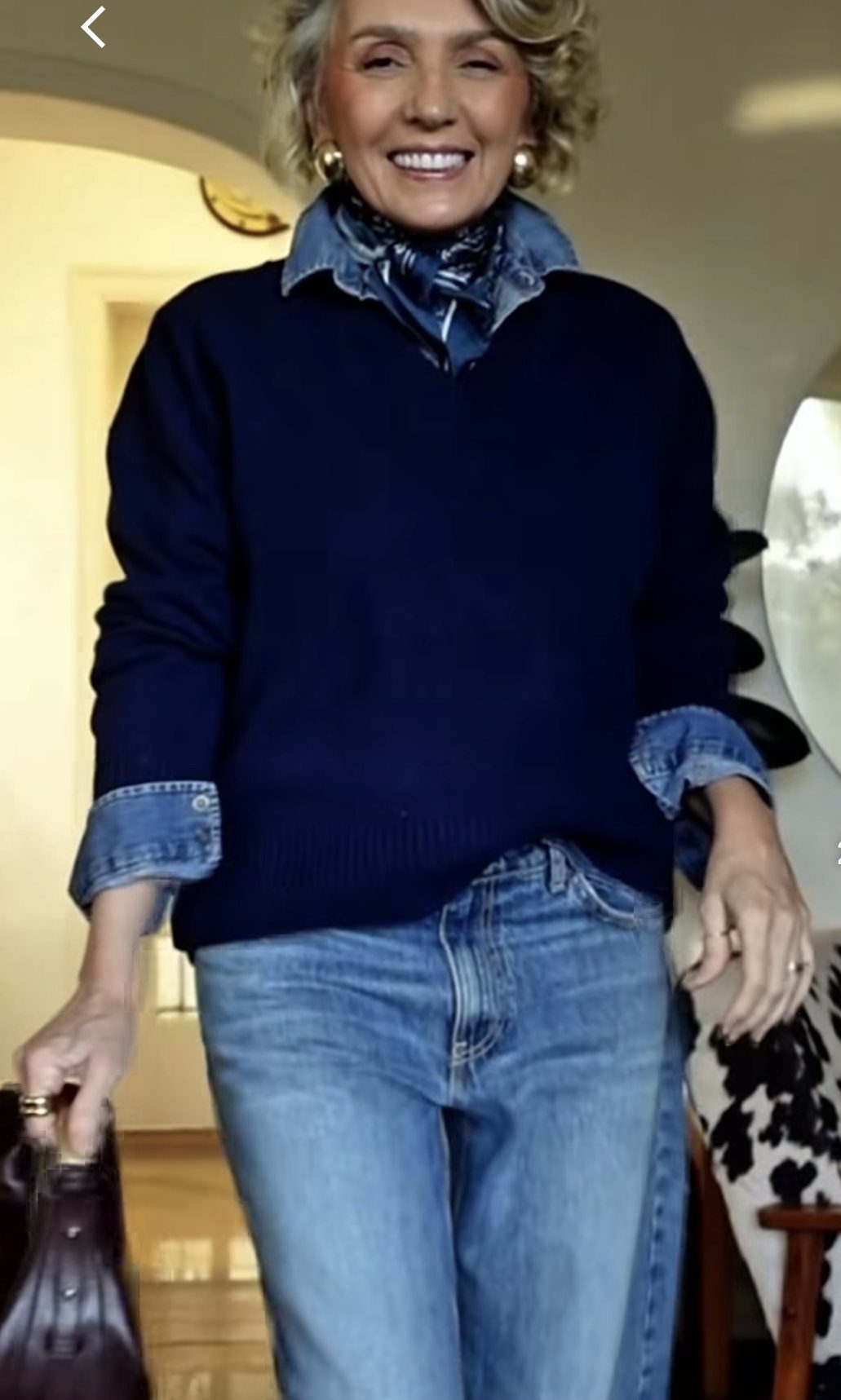 Smiling woman wearing a navy sweater layered over a denim shirt and blue jeans.