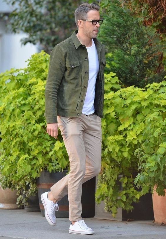 A man walking in an olive suede jacket, white t-shirt, beige chinos, and white sneakers.