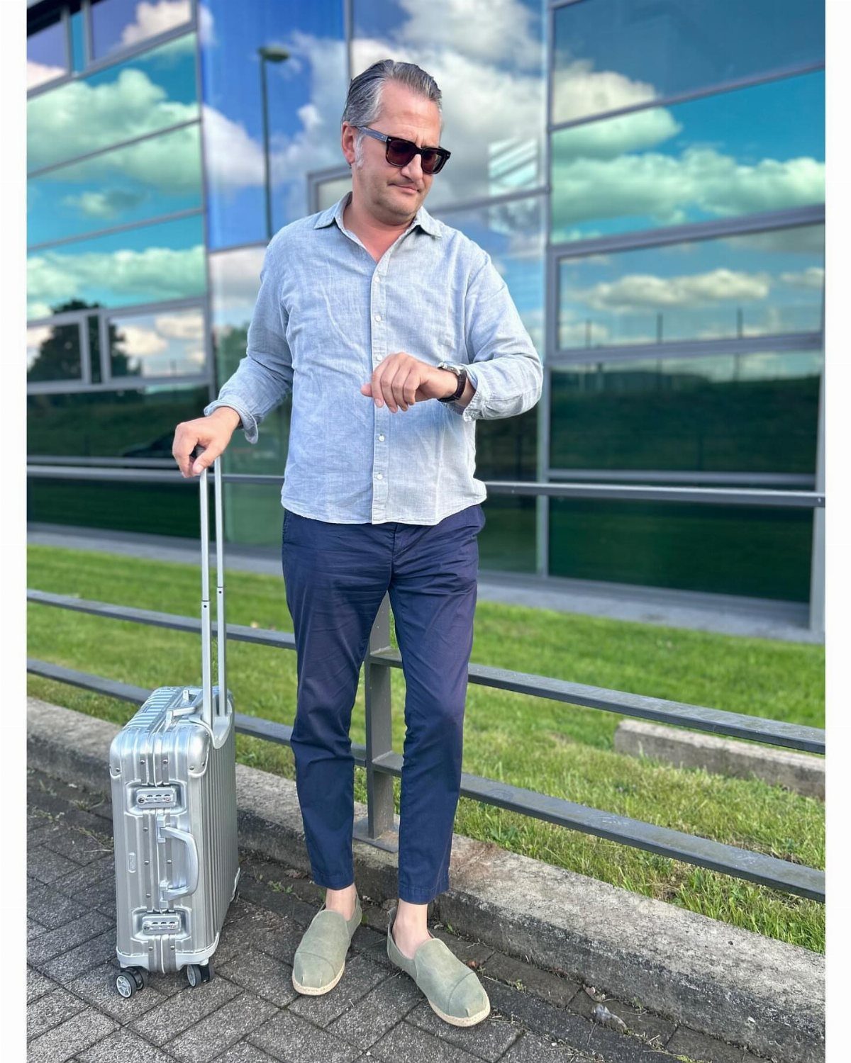 Man in smart casual attire standing with a silver suitcase outdoors near a glass building.