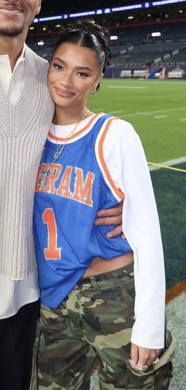 A woman wearing a blue basketball jersey over a white long-sleeve shirt and camouflage pants.