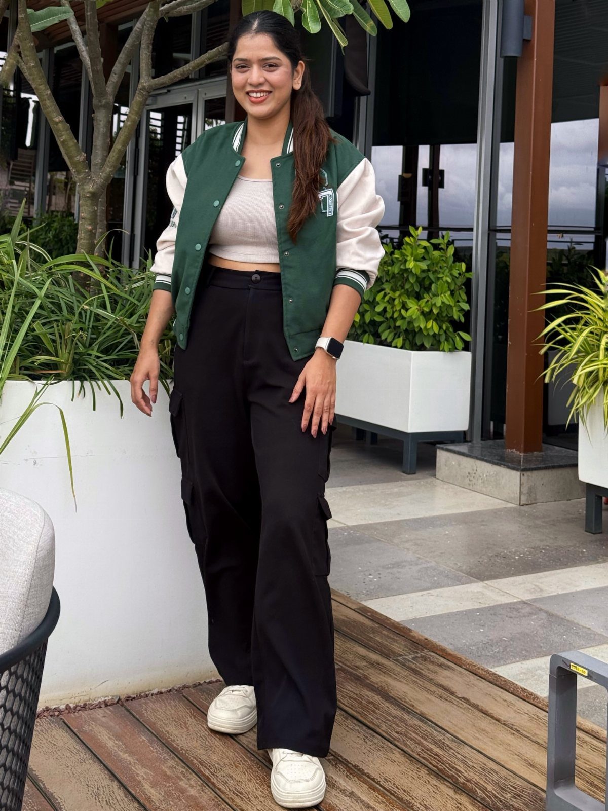 Woman wearing a green varsity jacket, beige top, black cargo pants, and white sneakers.