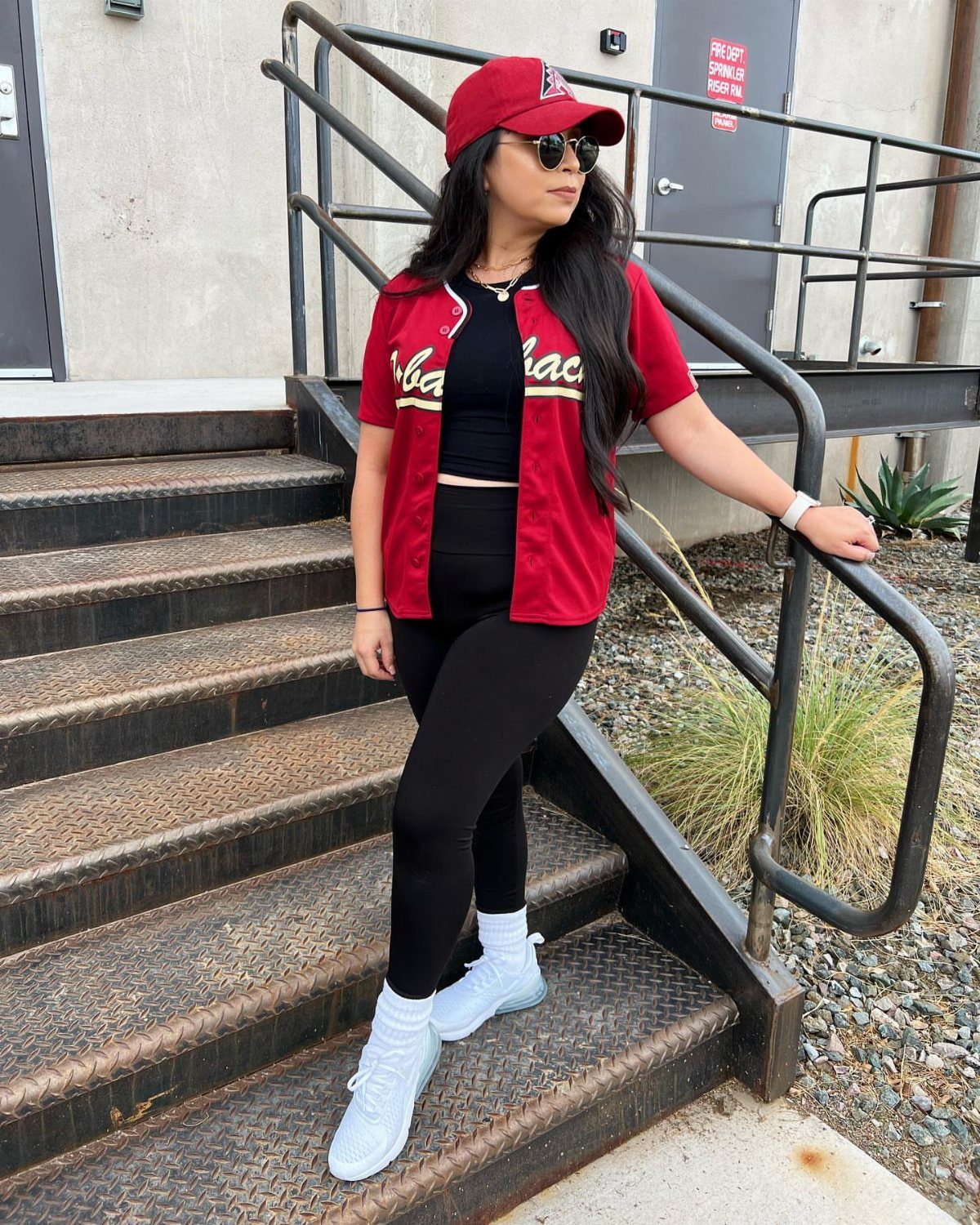 Woman in a red baseball jersey, black leggings, and white sneakers posing on stairs.