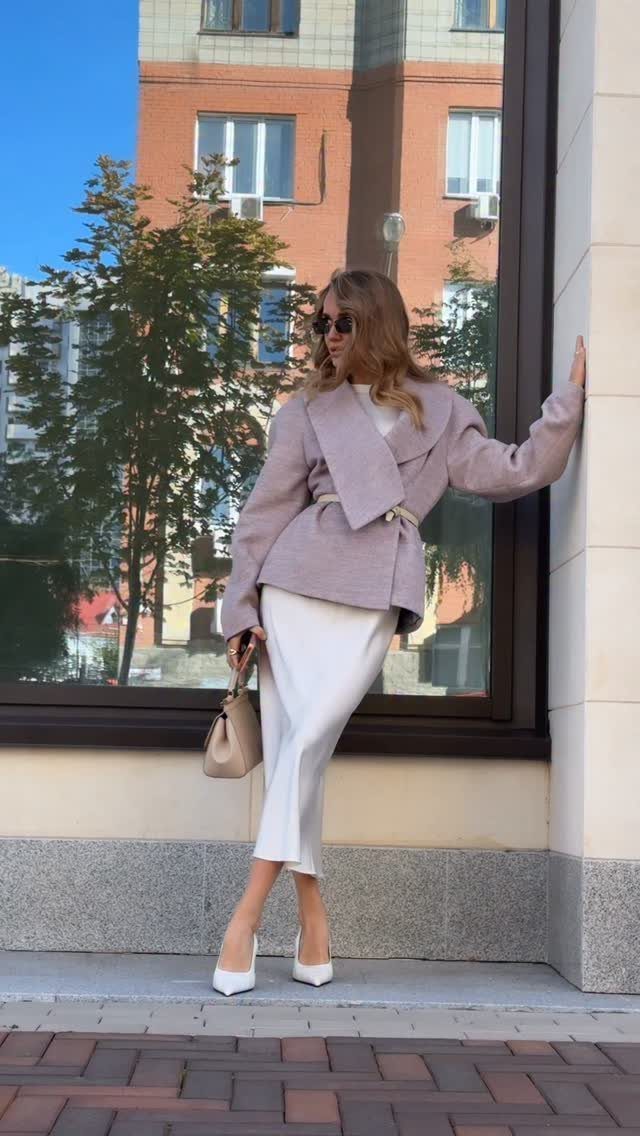 Woman in a white midi skirt and lavender blazer posing against a glass building window.
