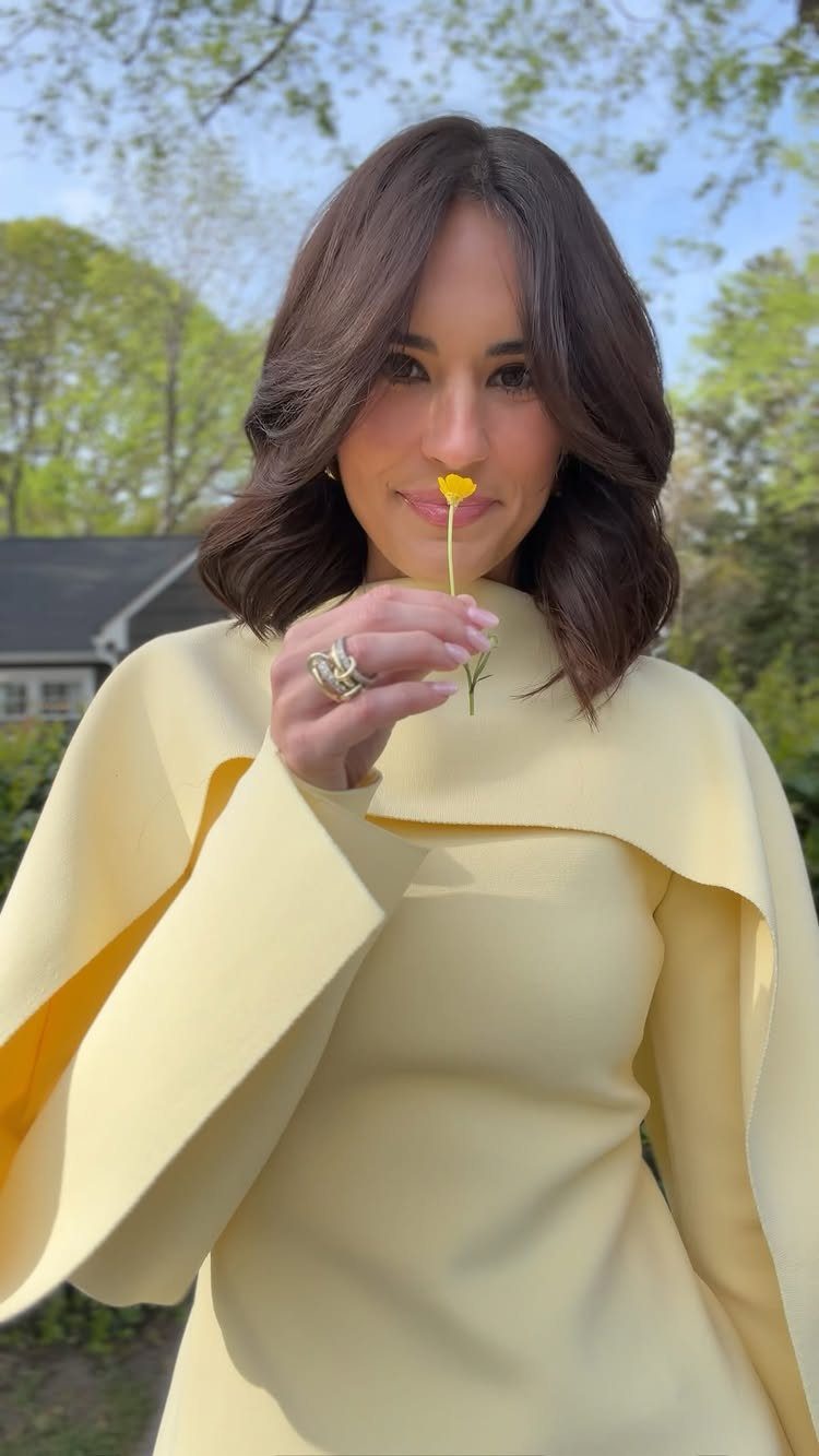 Woman in a pale yellow top holding a small yellow flower in a garden setting.