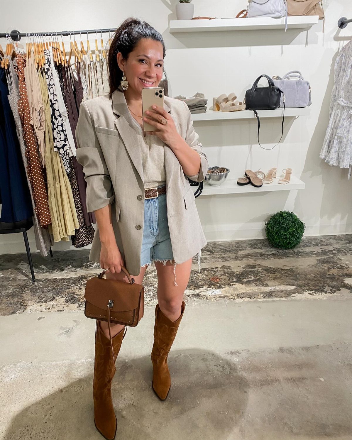 Smiling woman in a beige blazer and light blue denim shorts posing for a mirror selfie.