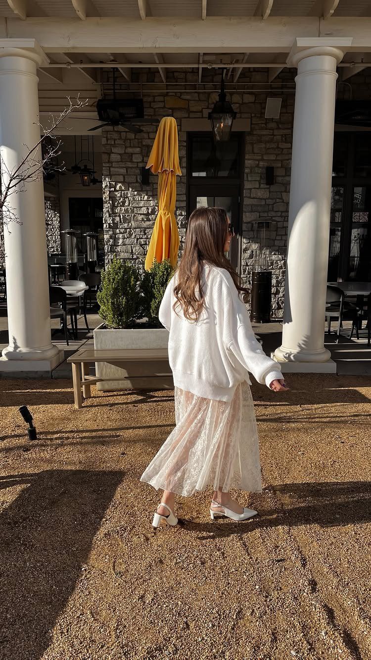 Woman walking in a white sweater and a cream lace midi skirt outdoors.