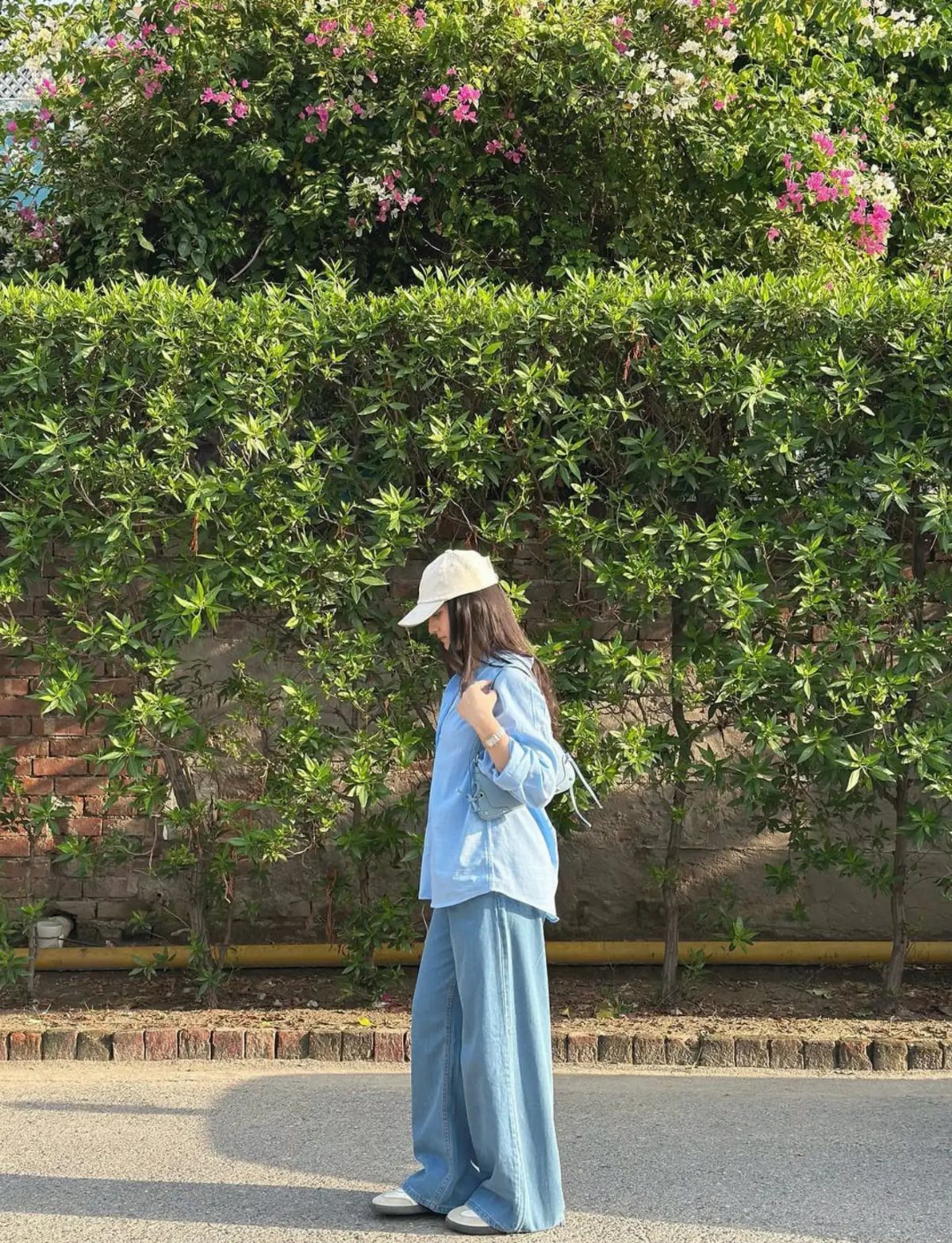 Woman in light blue shirt and wide-leg denim trousers standing by a green hedge.