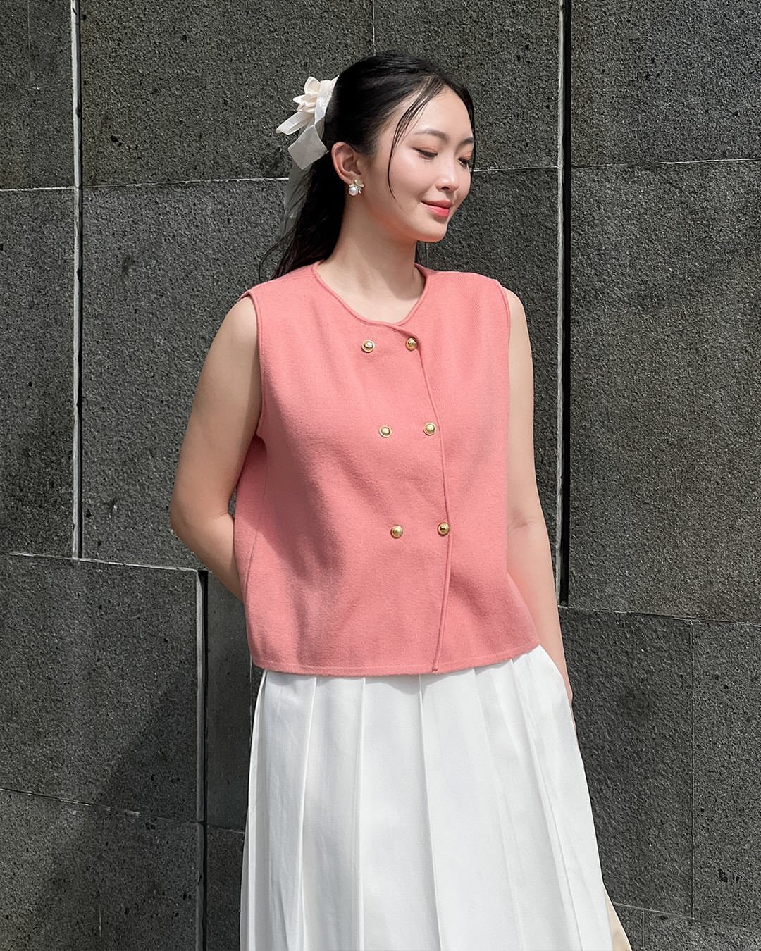 Woman wearing a coral sleeveless structured top with gold buttons and a white pleated skirt.