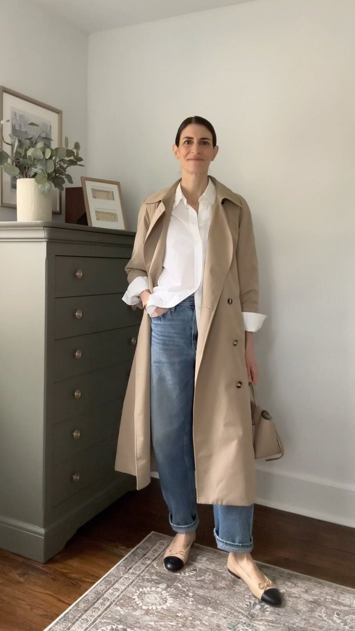 Woman wearing a long beige trench coat, white shirt, blue jeans, and black ballet flats.