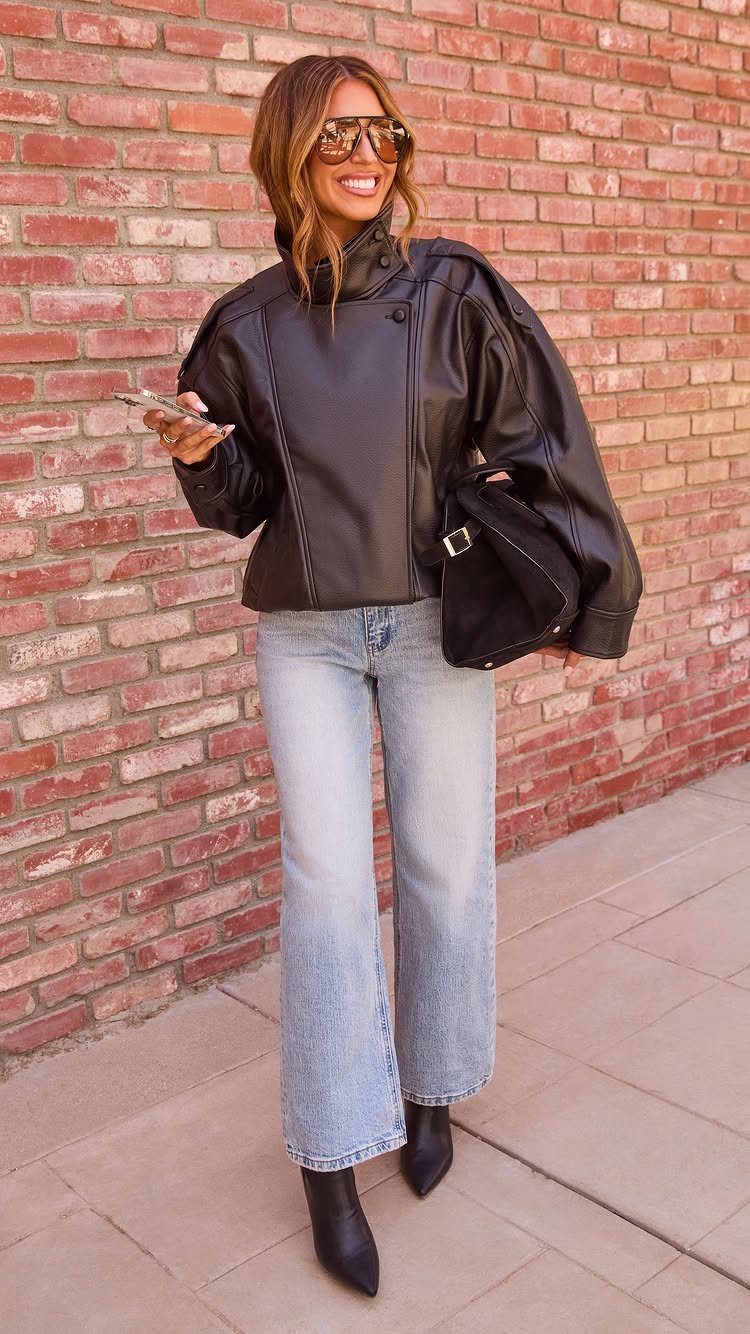 A woman posing in a black faux leather jacket, light blue jeans, and black boots.
