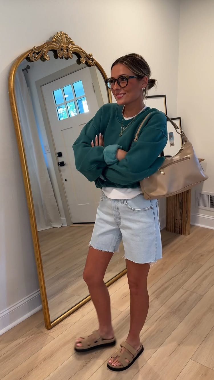A woman posing in a teal oversized sweatshirt and denim shorts by a mirror.