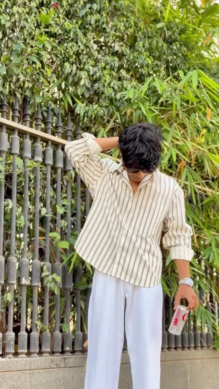 A person wearing a striped shirt and white relaxed pants standing by a green hedge.
