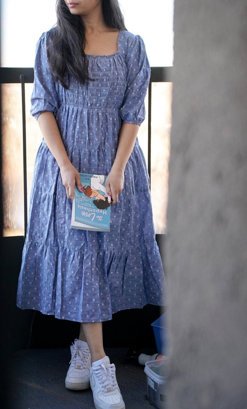 Woman wearing a blue patterned midi dress and white sneakers while holding a book.