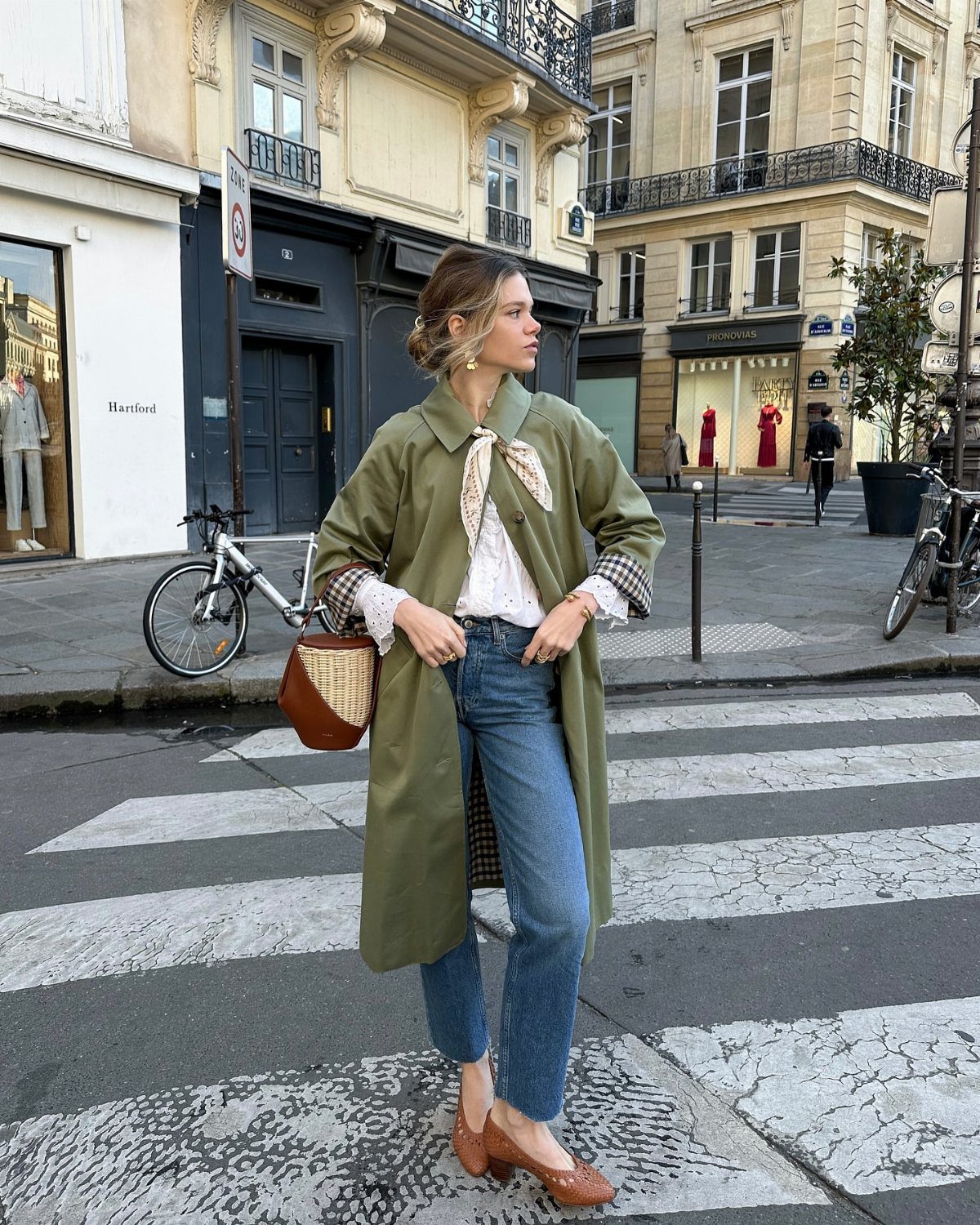 A woman wearing an olive green trench coat, white blouse, and blue jeans standing on a city street.