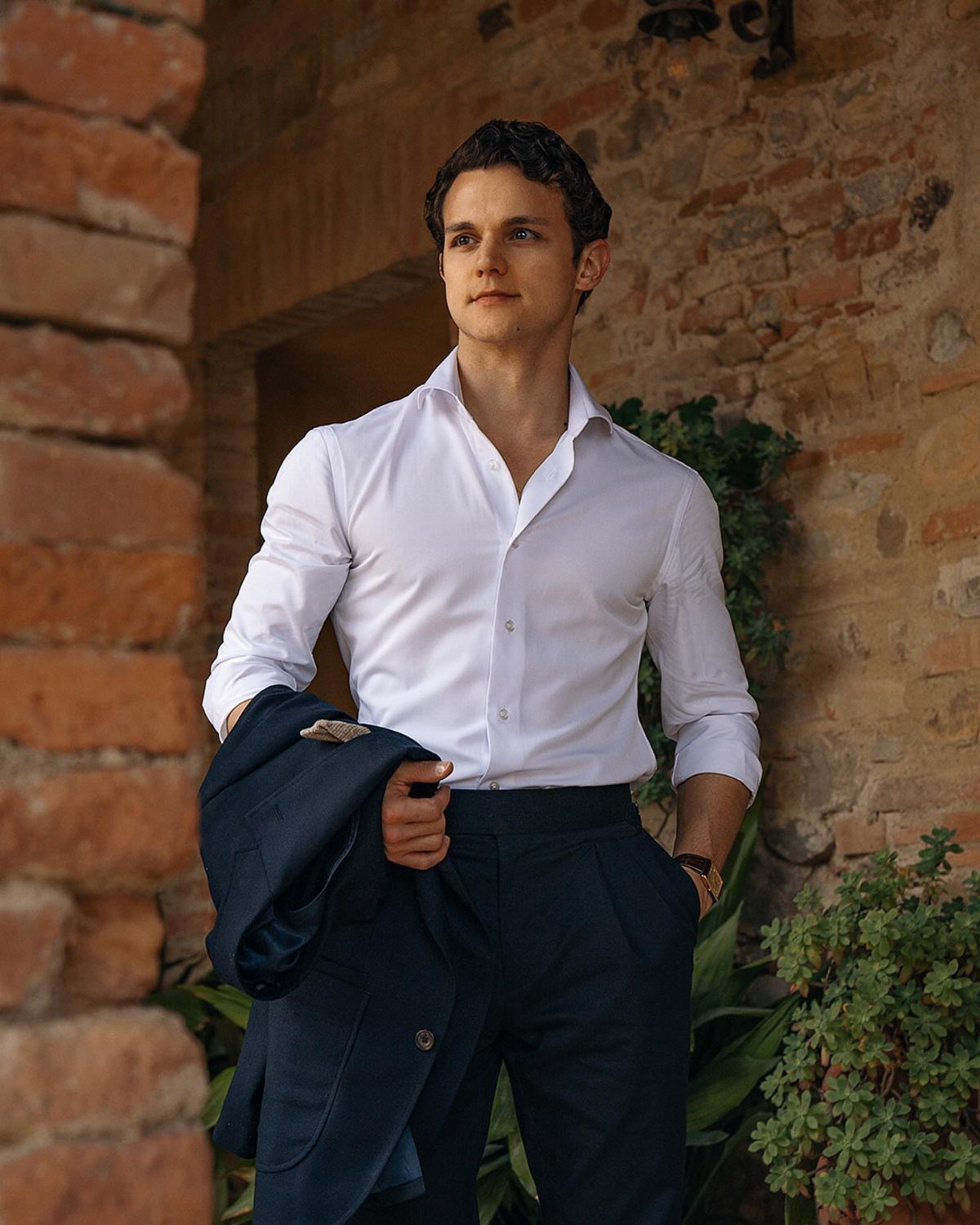 Man in a white button-down shirt and navy trousers standing by a brick wall.