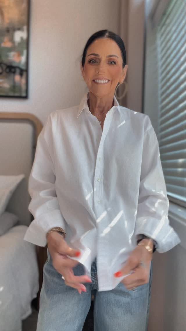 A woman smiling while wearing a white button-down shirt and light blue jeans.