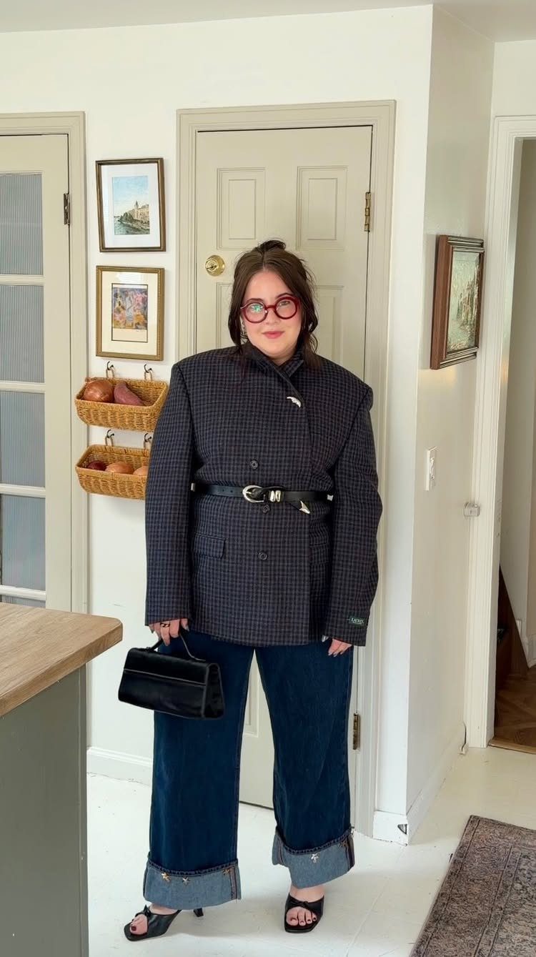 Woman wearing a belted patterned blazer, wide-leg jeans, and black heels standing in a hallway.