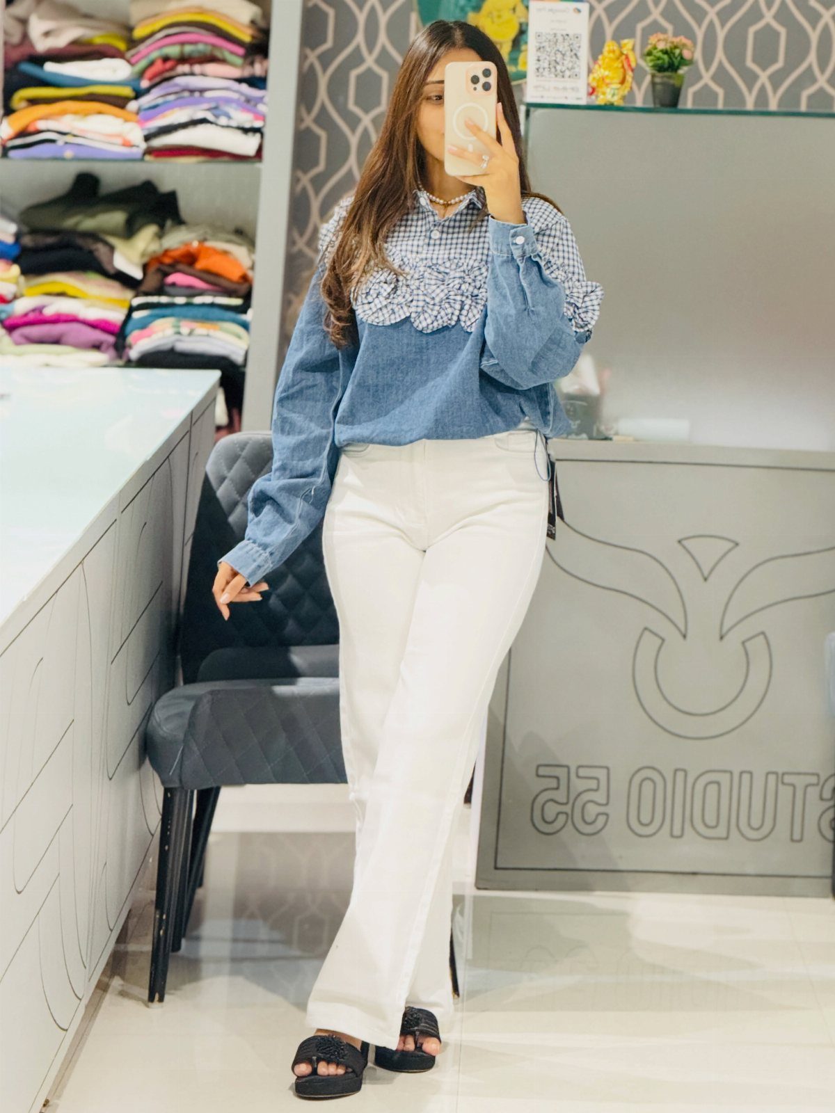 Woman in a blue ruffled statement top and white trousers taking a mirror selfie.