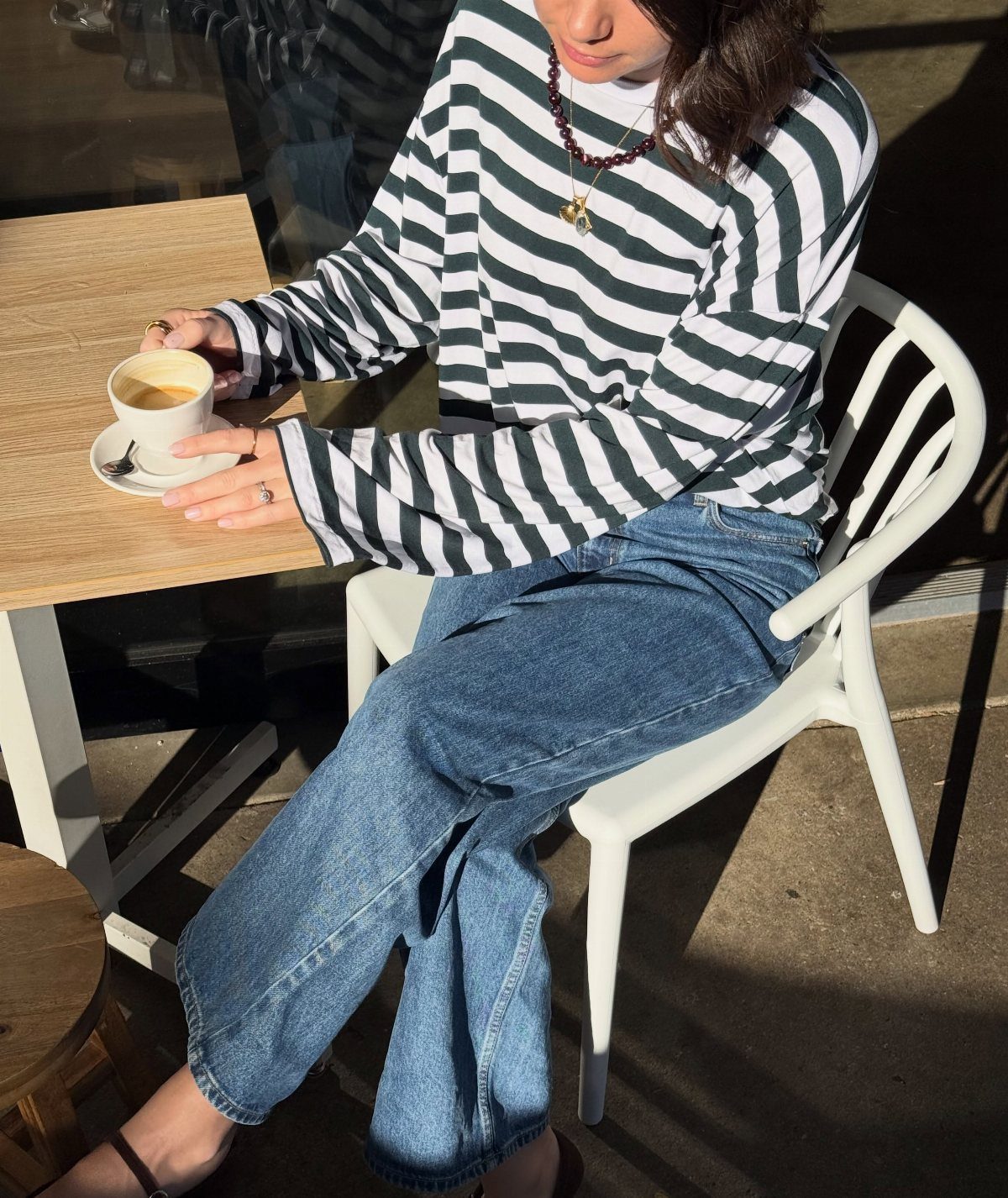 Person in striped shirt and blue jeans sitting at a cafe table with coffee.