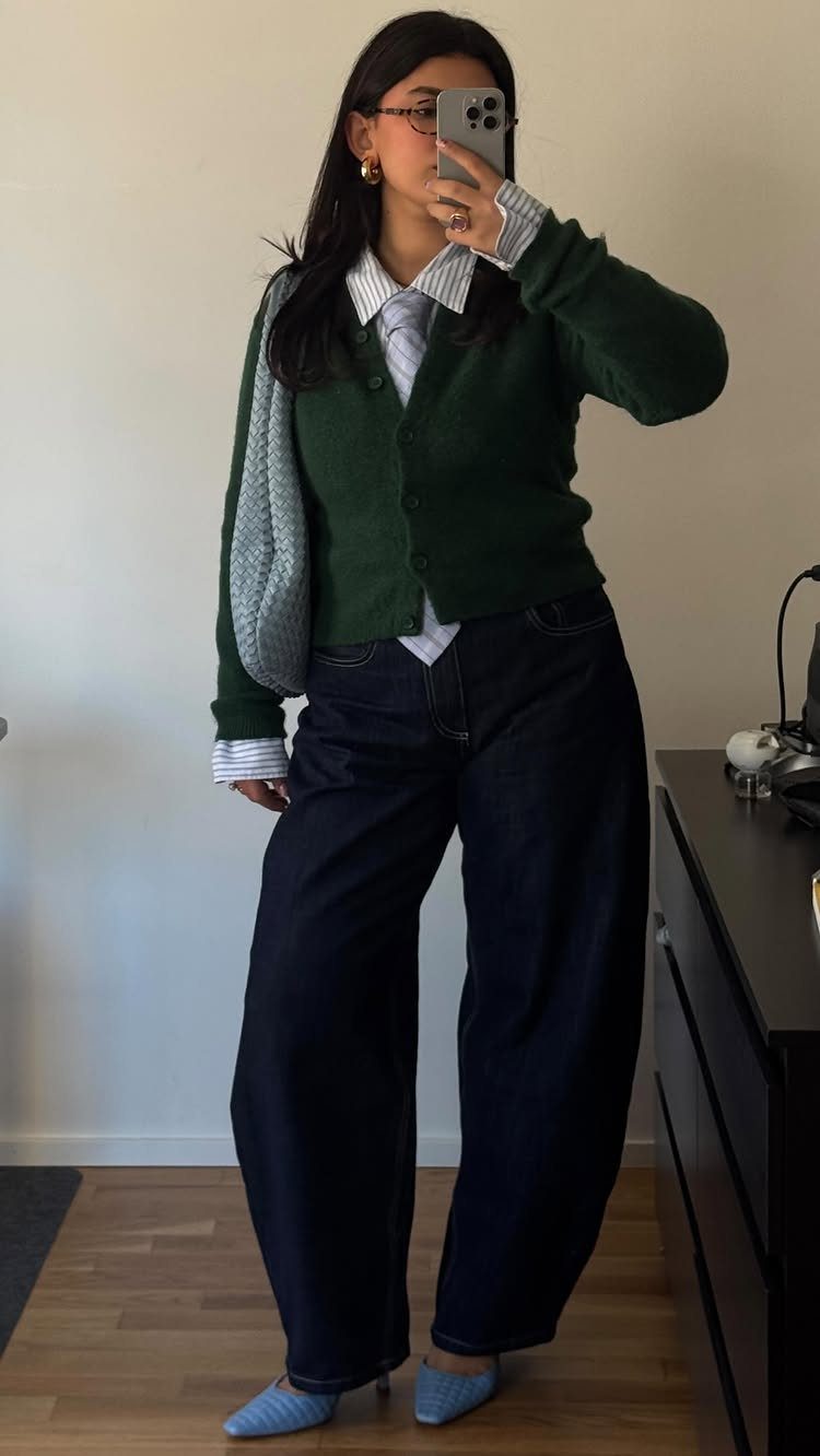 A woman posing in a green cardigan, striped shirt, wide-leg jeans, and blue flats.