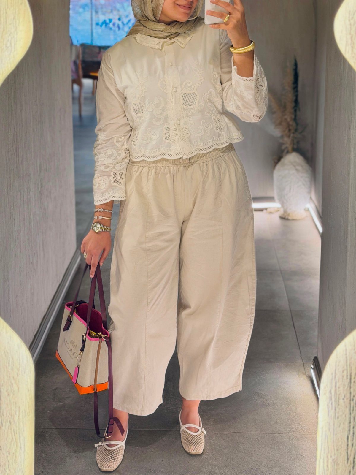 Woman wearing a white lace blouse and beige wide-leg cropped trousers holding a tote bag.