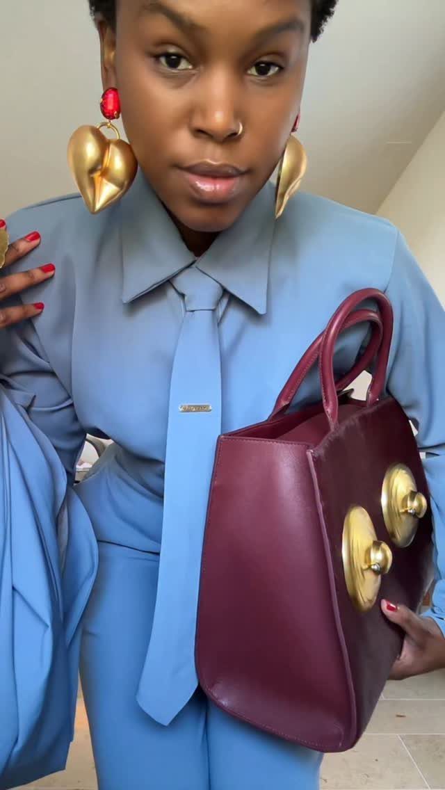 Person wearing a light blue monochromatic shirt, tie, and trousers while holding a burgundy handbag.