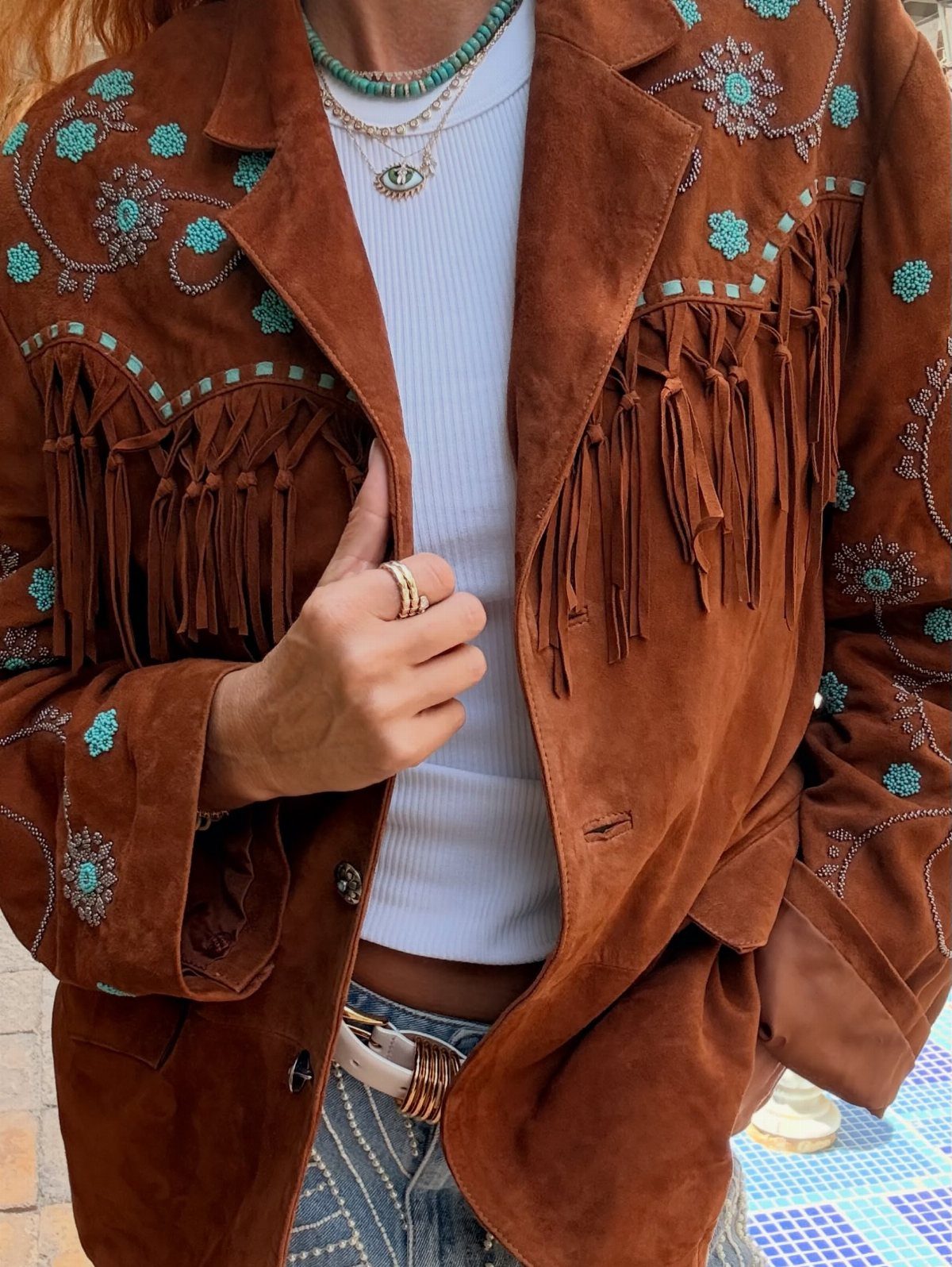 A person wearing a brown suede jacket with turquoise embroidery and long fringe detailing.