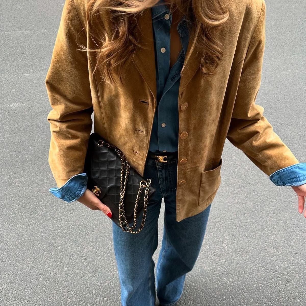 A person wearing a tan suede jacket, denim shirt, blue jeans, and carrying a black quilted handbag.