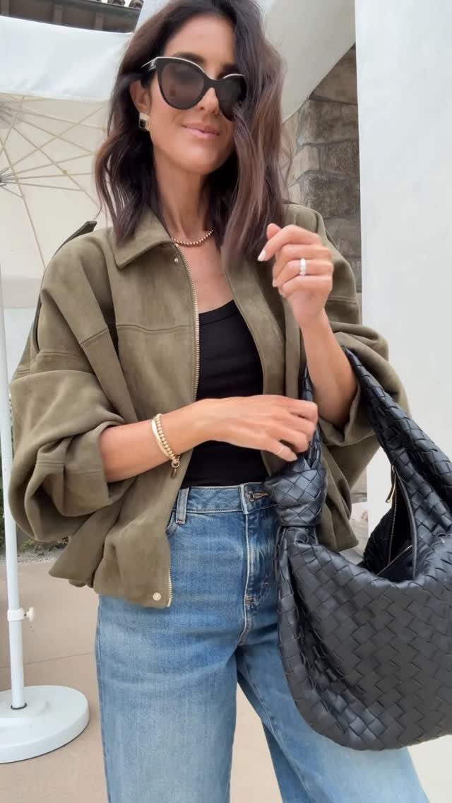 Woman wearing an olive green suede bomber jacket, black top, and blue jeans outdoors.