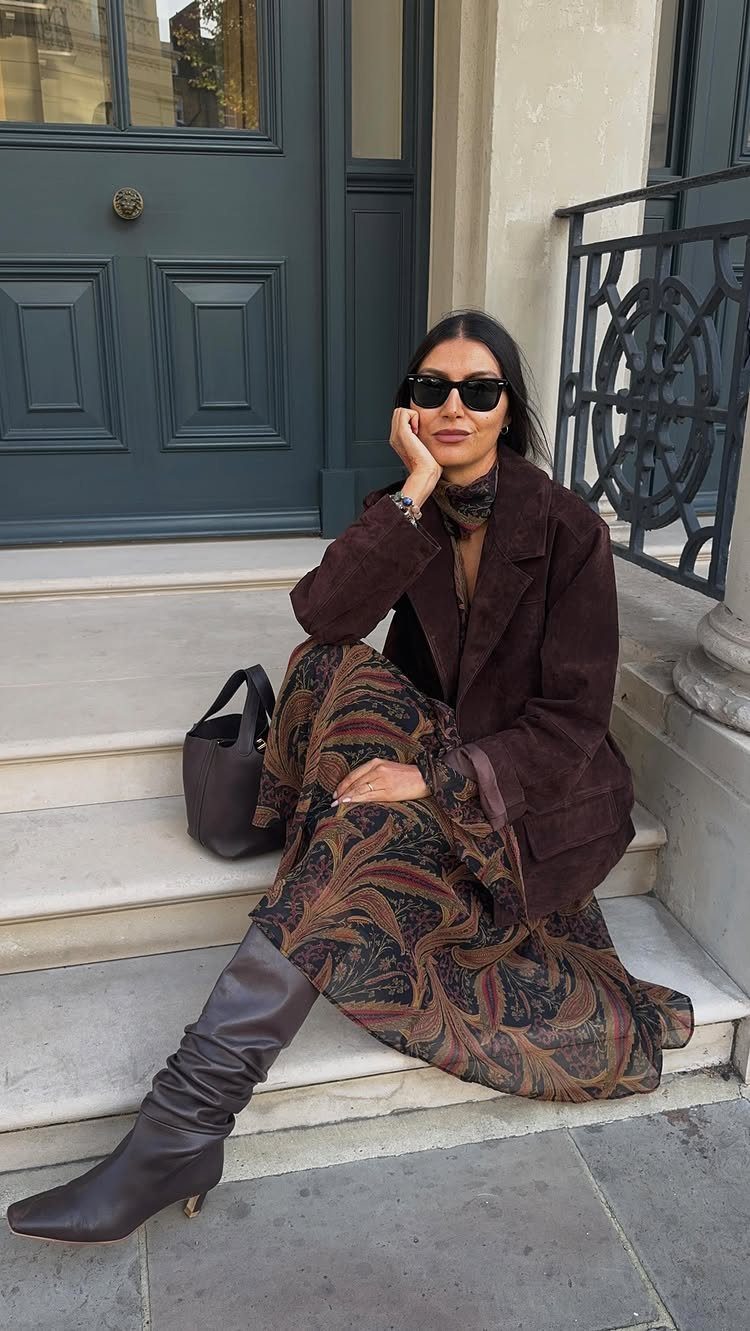 Woman in a brown suede jacket and patterned maxi dress sitting on outdoor steps.