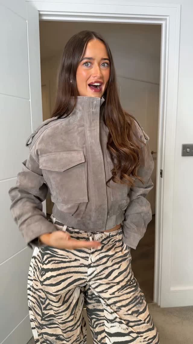 A woman wearing a grey suede jacket and zebra-print trousers standing in a doorway.