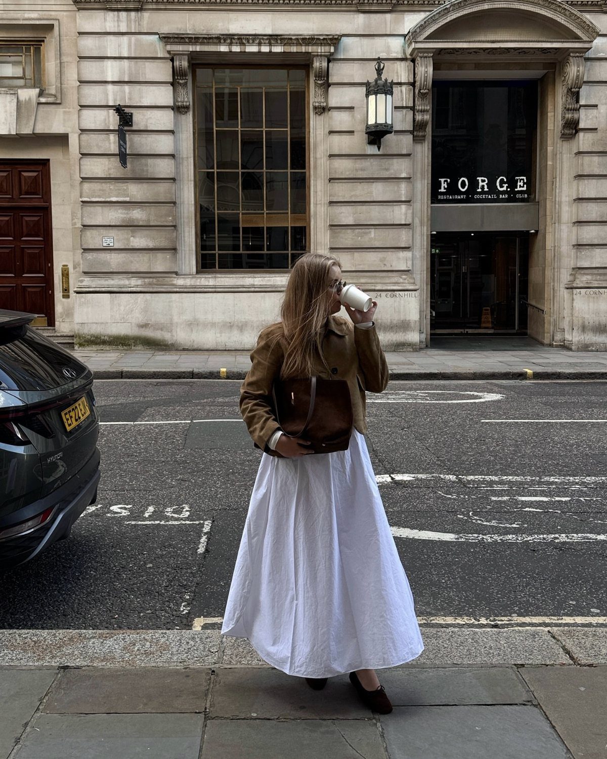 Woman in a brown suede jacket and long white skirt standing on a city street.