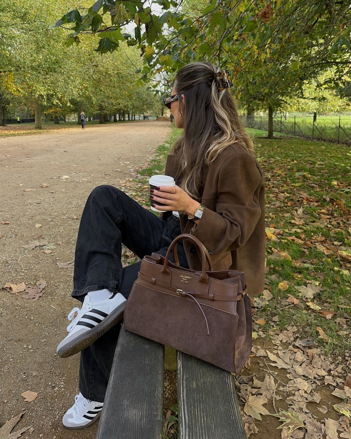 Woman in a brown suede jacket and black jeans sitting on a park bench.