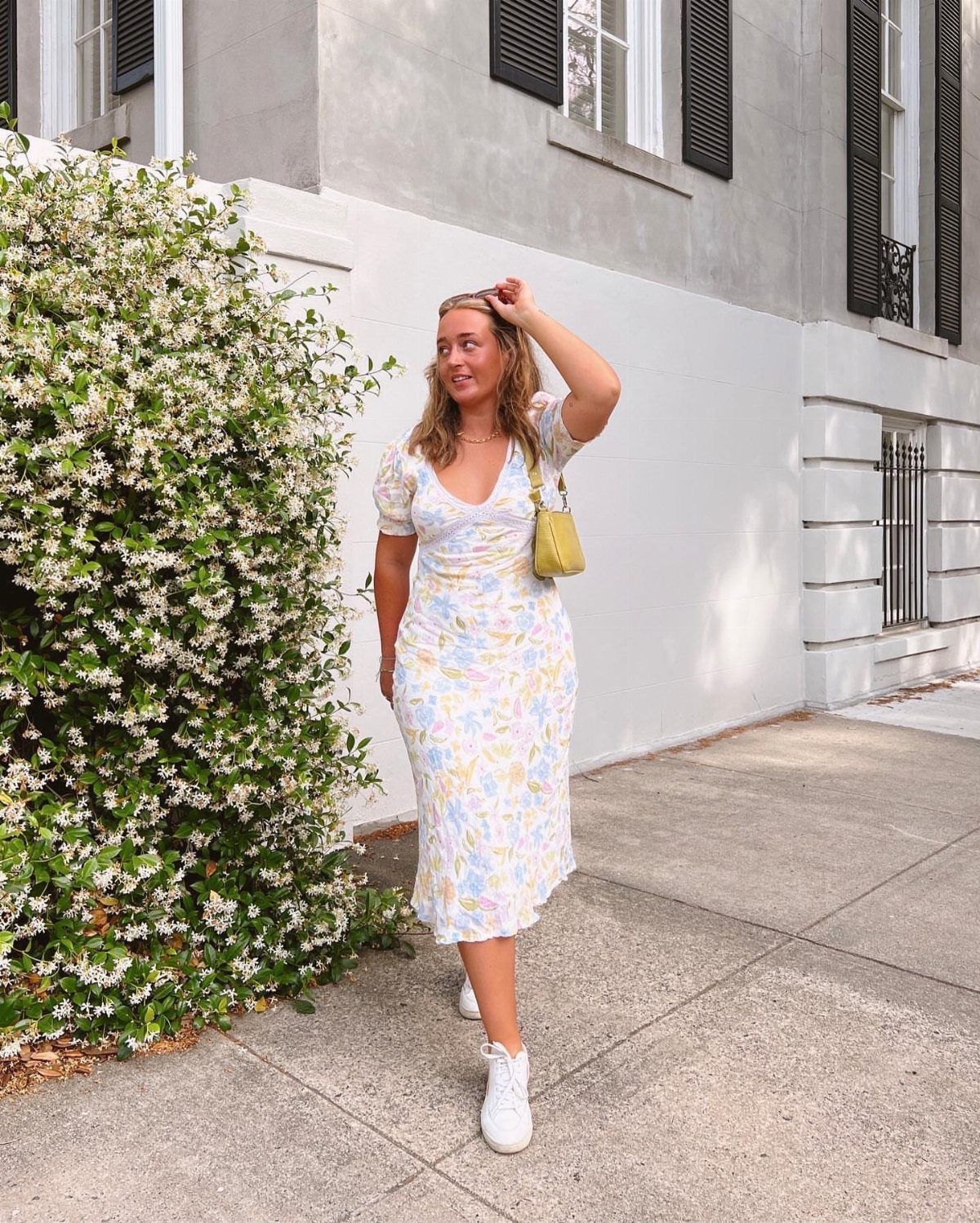 A woman in a floral midi dress and white sneakers standing by a flowering bush.