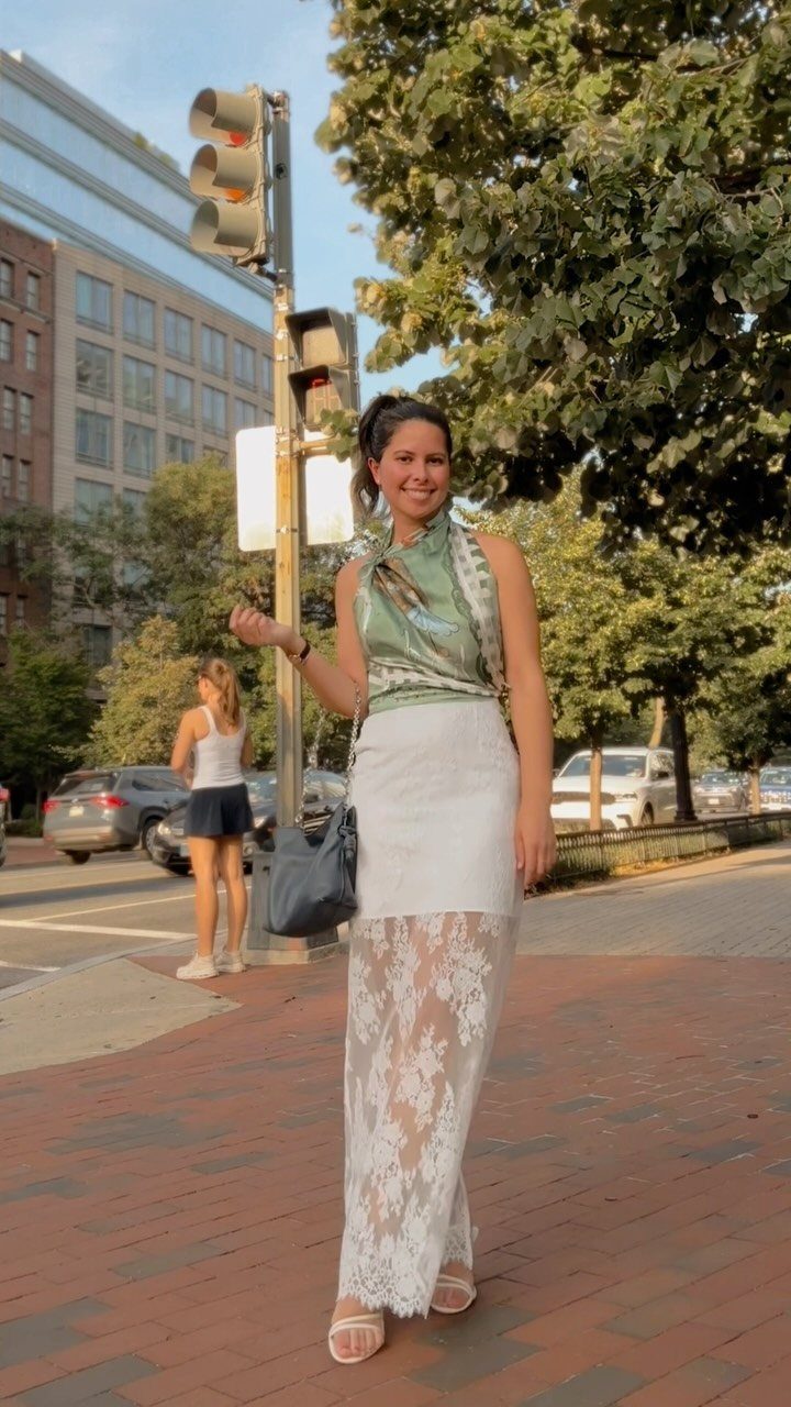 Smiling woman in a green halter top and white lace skirt standing on a city sidewalk.