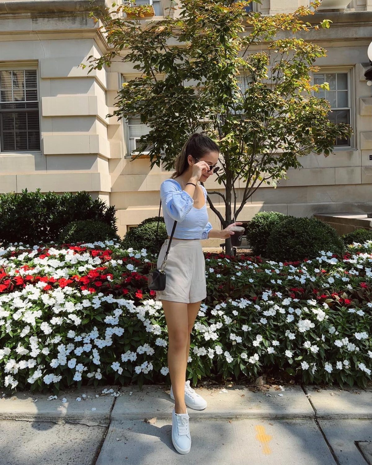 Woman wearing a light blue off-the-shoulder crop top and beige high-rise shorts standing by flowers.