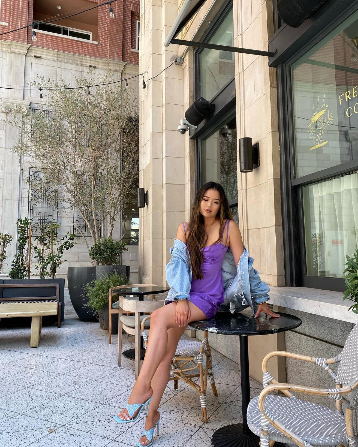 Woman in a purple satin mini dress and denim jacket sitting at an outdoor cafe table.