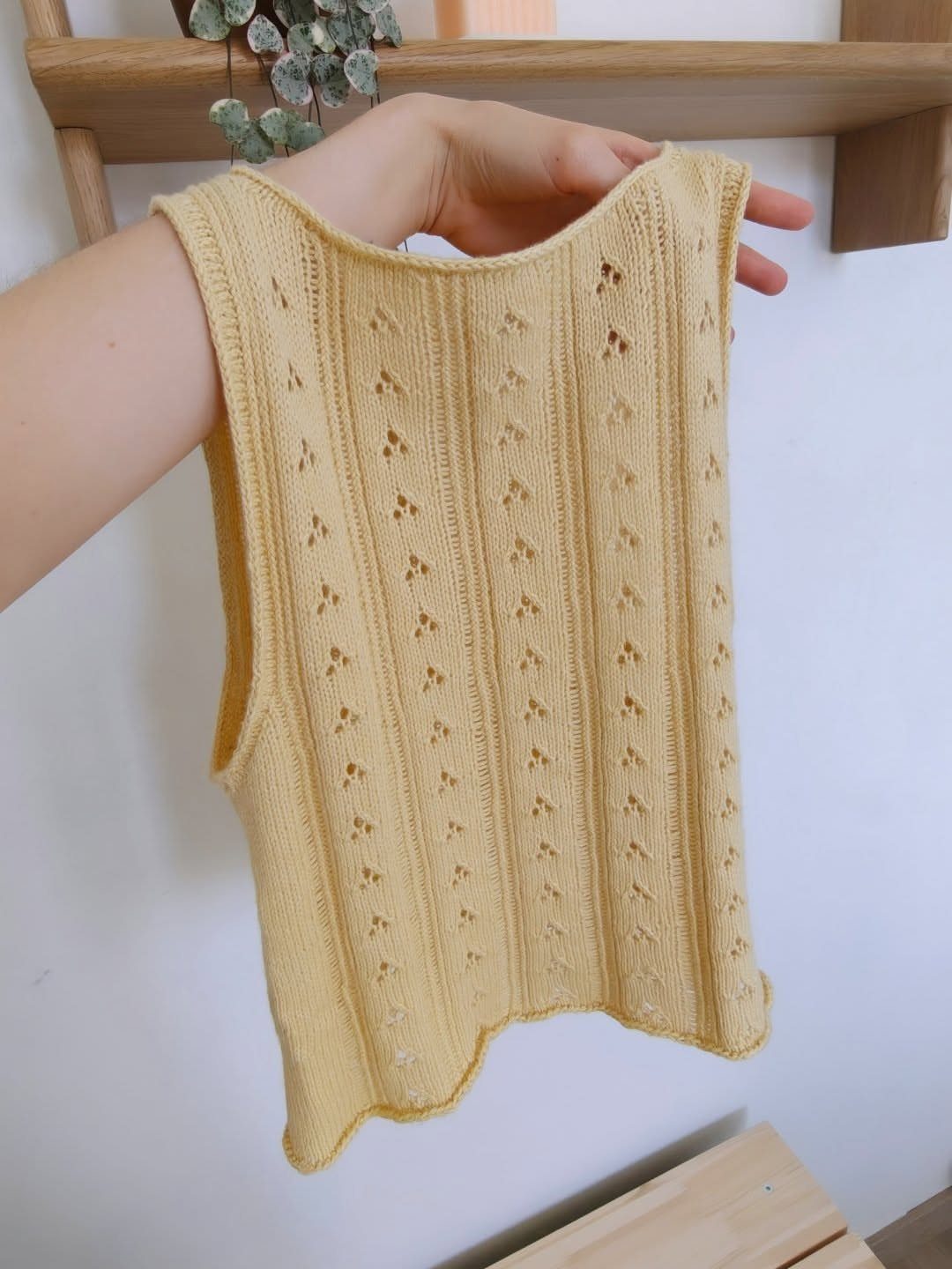 A hand holds a pale yellow sleeveless knit top with eyelet lace details against a white wall.
