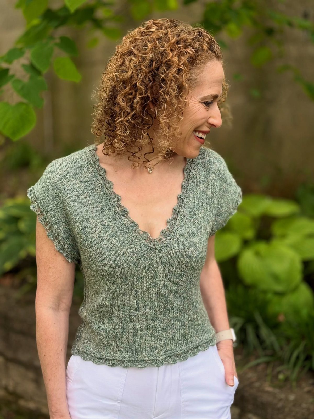 A smiling woman with curly hair wearing a light green textured knit short-sleeve top outdoors.