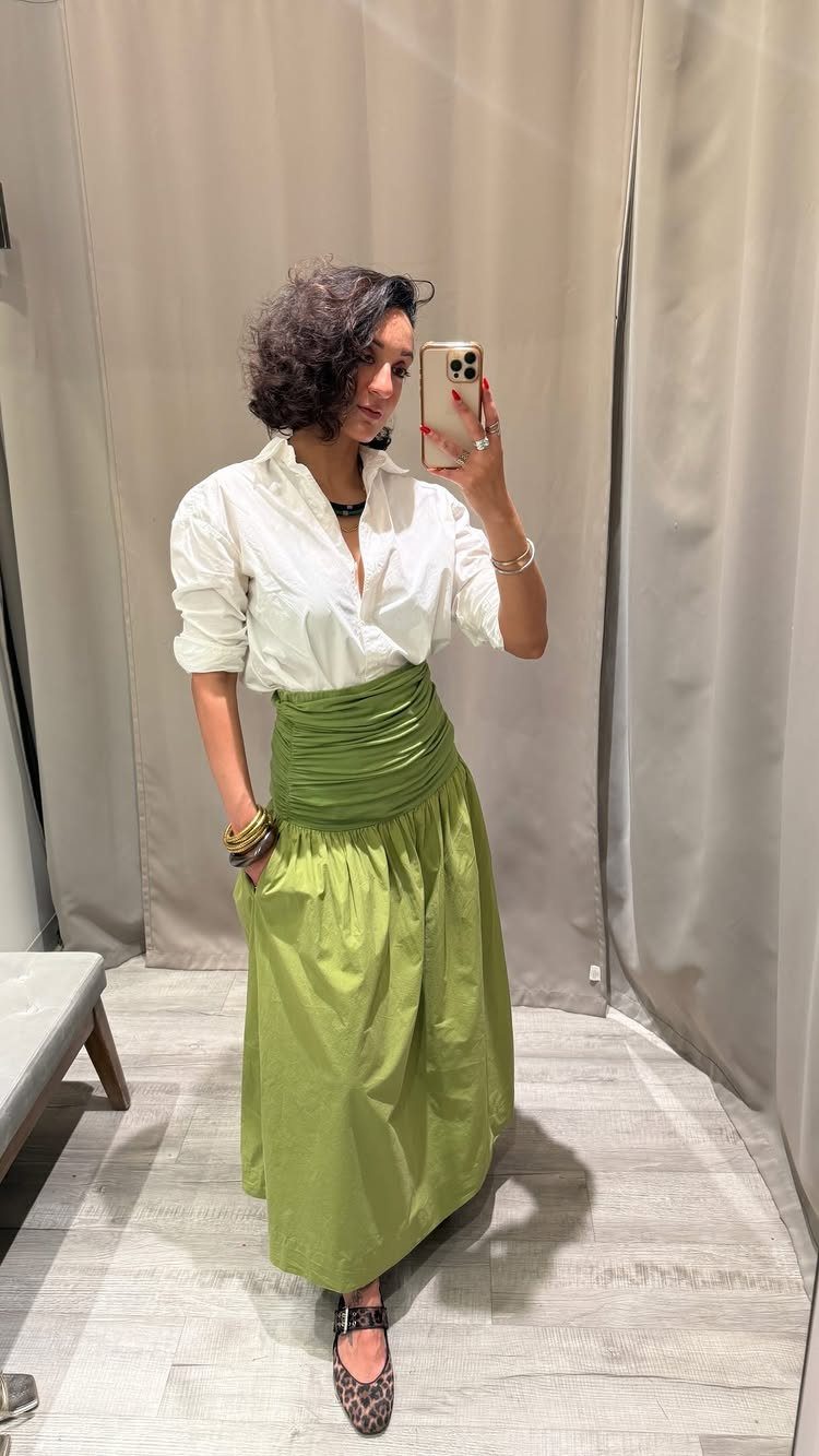 Woman in a white shirt and long olive green skirt posing in a fitting room.