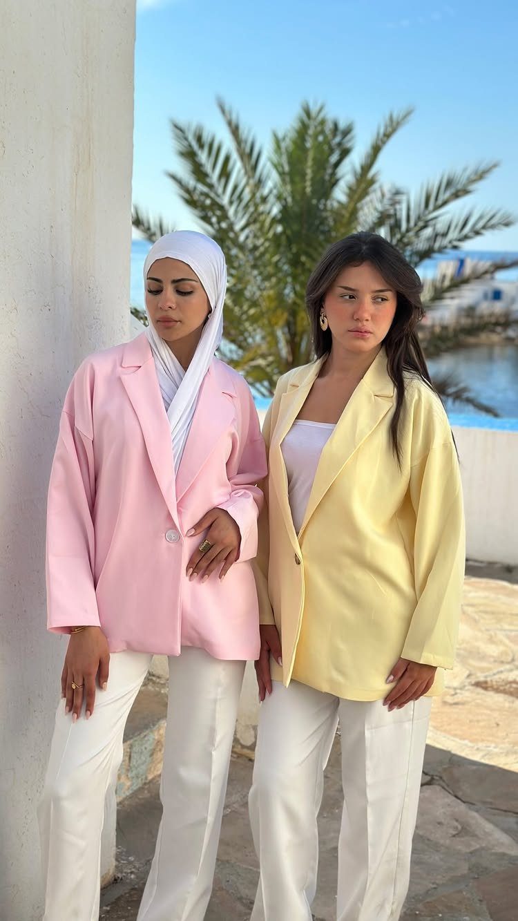 Two women posing in pastel blazers and white trousers against a tropical outdoor background.