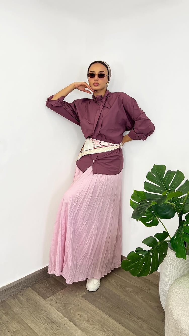 Woman wearing a mauve shirt and light pink pleated maxi skirt with a patterned waist scarf.