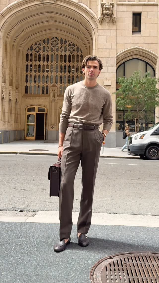A man in a beige sweater and brown trousers holding a briefcase on a city street.