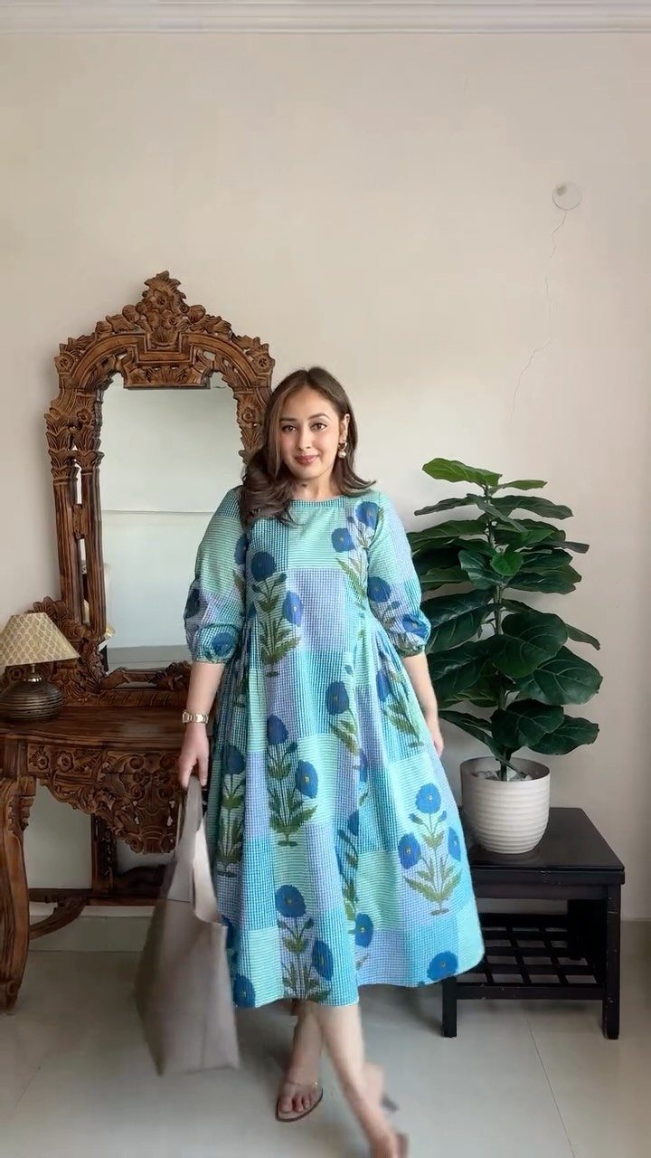 A woman posing in a blue patterned summer dress holding a large tote bag.