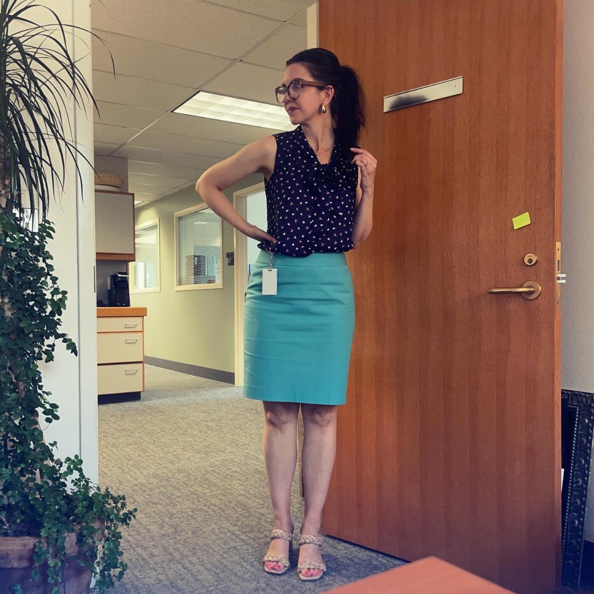 A woman posing in an office wearing a polka-dot top and a turquoise pencil skirt.